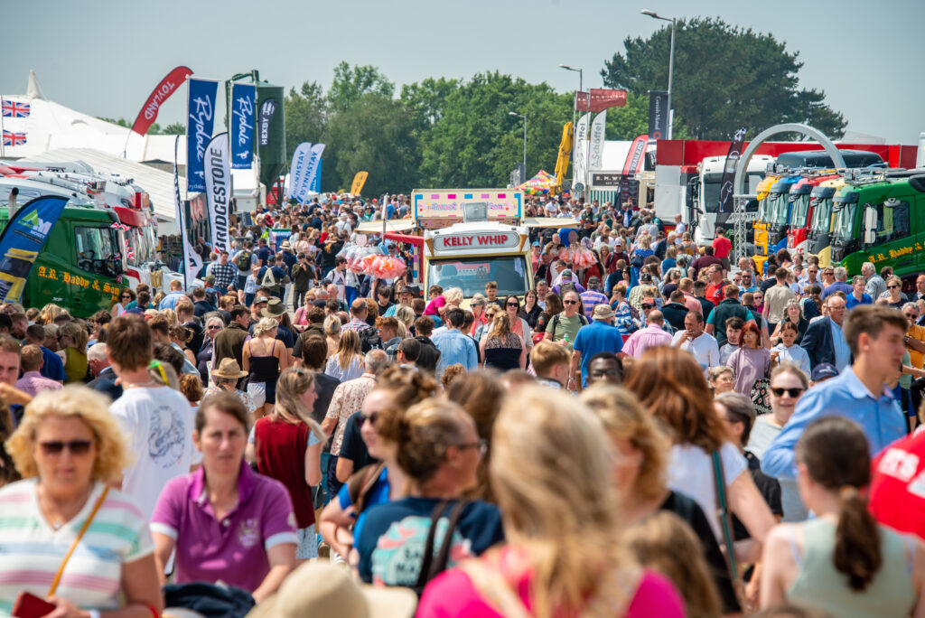Hundreds of people in a huge crowd walk between stalls, flags and vans at the Royal Cornwall Show 2024. There is one single ice cream van in the middle of all the hustle and bustle