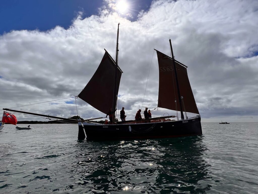 A classic sailing boat with two sails is cast in shadow against a moody, cloudy sky out on the water past Mousehole harbour in Cornwall. Expect scenes like this at the Sea Sail and Salts 2024 festival. There are three people in silhouette on the vessel