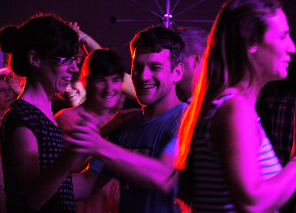 A couple dances in vibrant pink lighting during a ceilidh at the Lowender 2023 Cornish festival. There are other people dancing on the dance floor around them