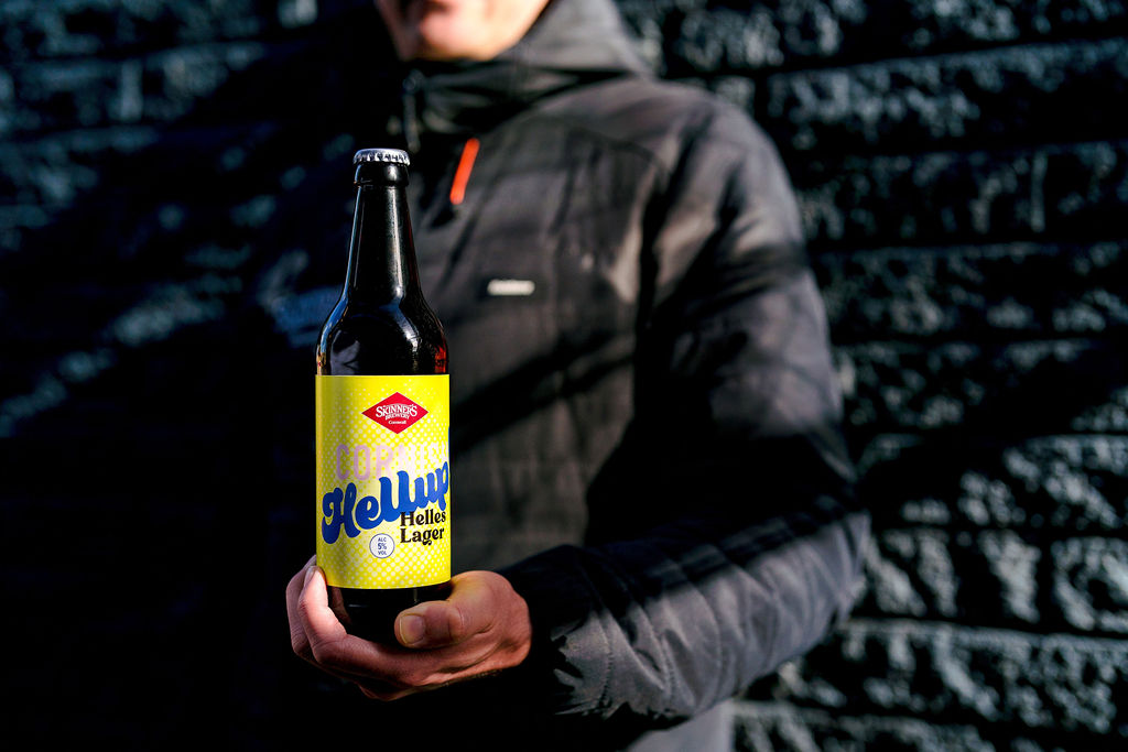 A man holds a bottle of Cornish Hellup! Helles Style lager in his hand. He stands against a black wall in a black coat