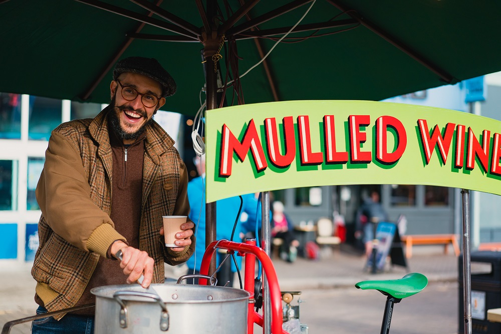 A mulled wine merchant with glasses and a moustache pours some of the festive drink into a cup at the Truro Christmas Market 2022. he smiles as he stands next to a sign saying 'Mulled Wine'