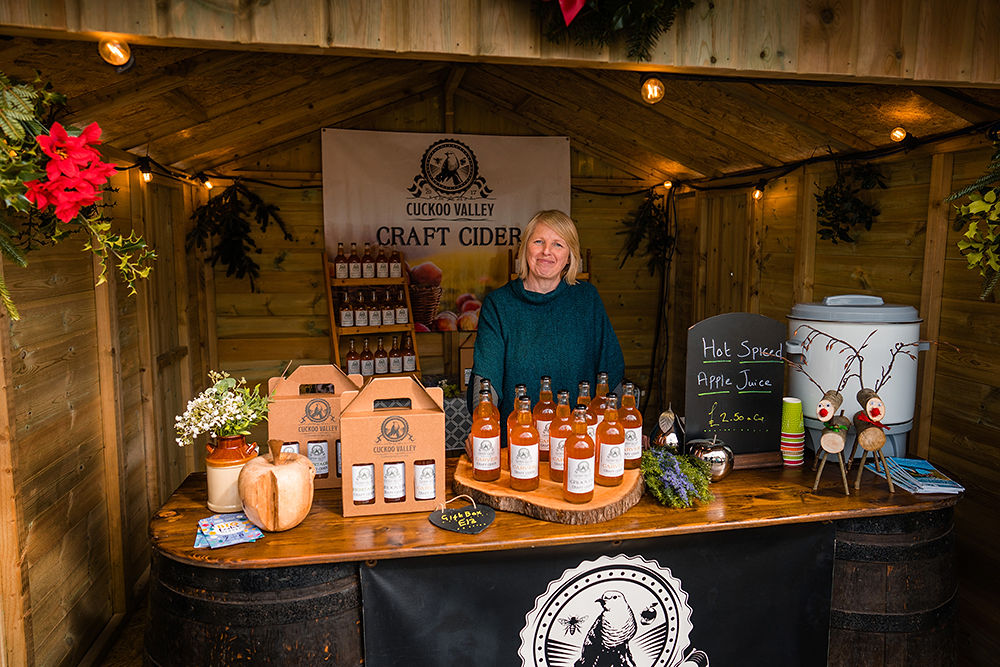 A woman smiles at us as she mans her Cuckoo Valley Ciders stall at the Truro Christmas Market 2023. The stall is festively lit