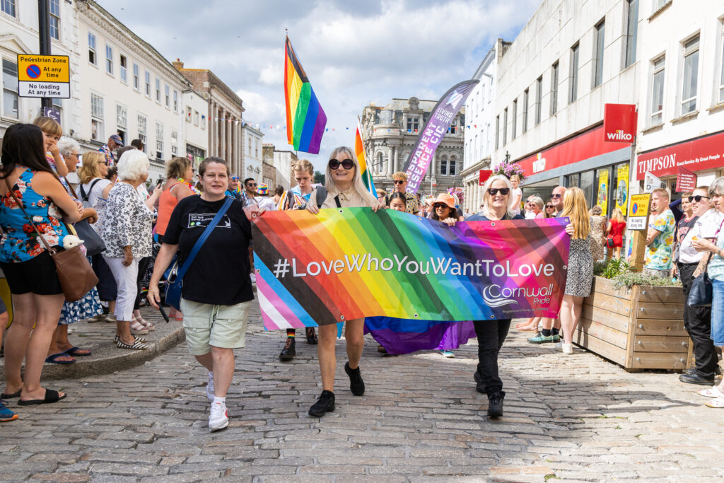 Cornwall Pride 2023 is going to be even bigger and better than Cornwall Pride 2022. Here, in 2022, people walk down a Cornish high street with rainbow flags and banners preaching love for all. Crowds of onlookers cheer this Pride parade.