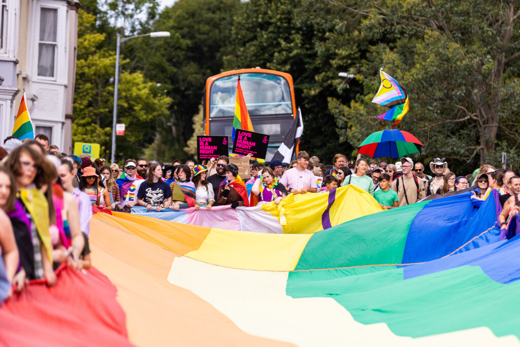 A huge rainbow flag is laid before a bus for Cornwall pride 2023. Hundreds of people hold it as the Cornwall Pride Bus stands and waits
