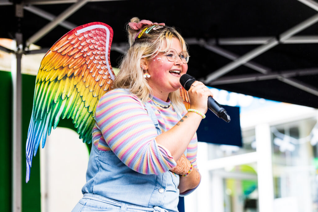 Pez the angel speaks to the crowds during Cornwall Pride 2023. She has a microphone in her hand, stands on a stage and has a set of rainbow-coloured angel wings on her back