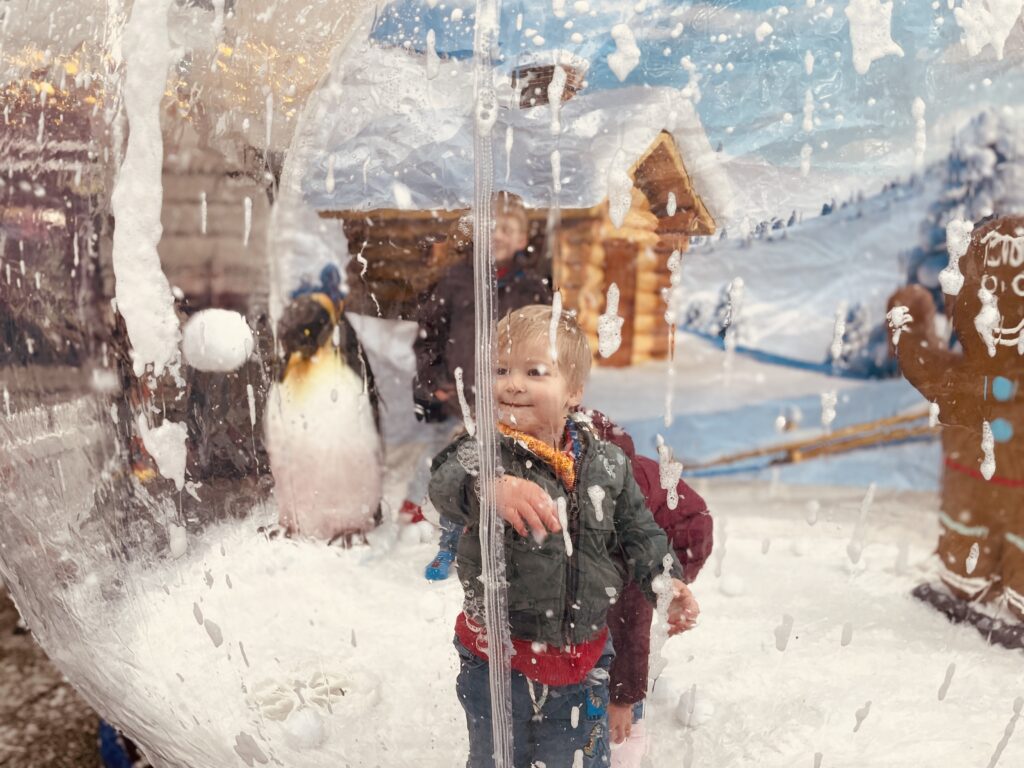 A little boy looks out from a giant snow globe in Falmouth for Falmouth's Festive Weekend 2023. There are penguins and a house covered in snow inside the big globe
