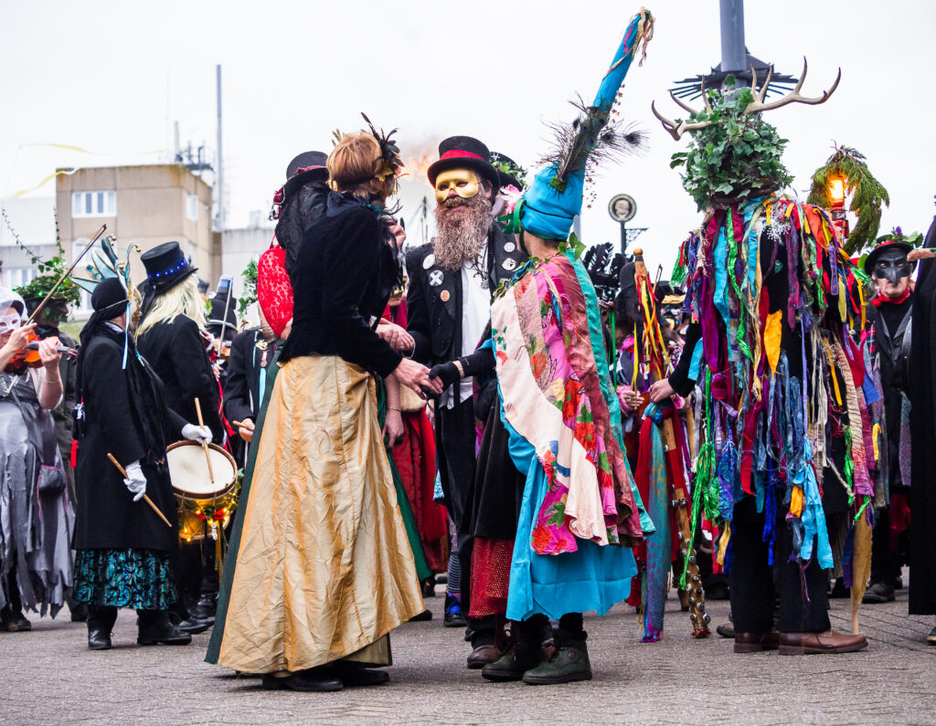 Two people in masks and bright costumes greet each other in a Penzance street for the Montol 2023 Festival. They are circled by many others in robes, masks and bright costumes