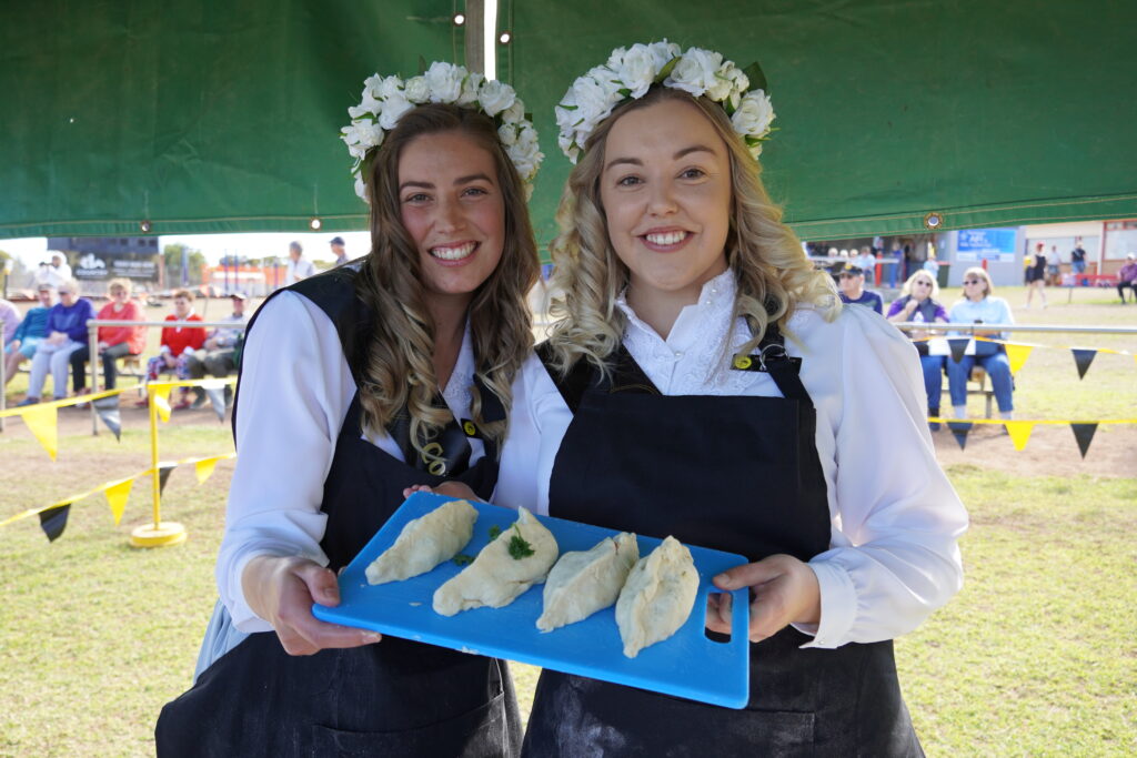 Two pretty girls in traditional Cornish clothes hold four pasties on a tray at the Kernewek Lowender 2023 festival in Australia. They have made them for the biggest Cornish festival outside of Cornwall in the world