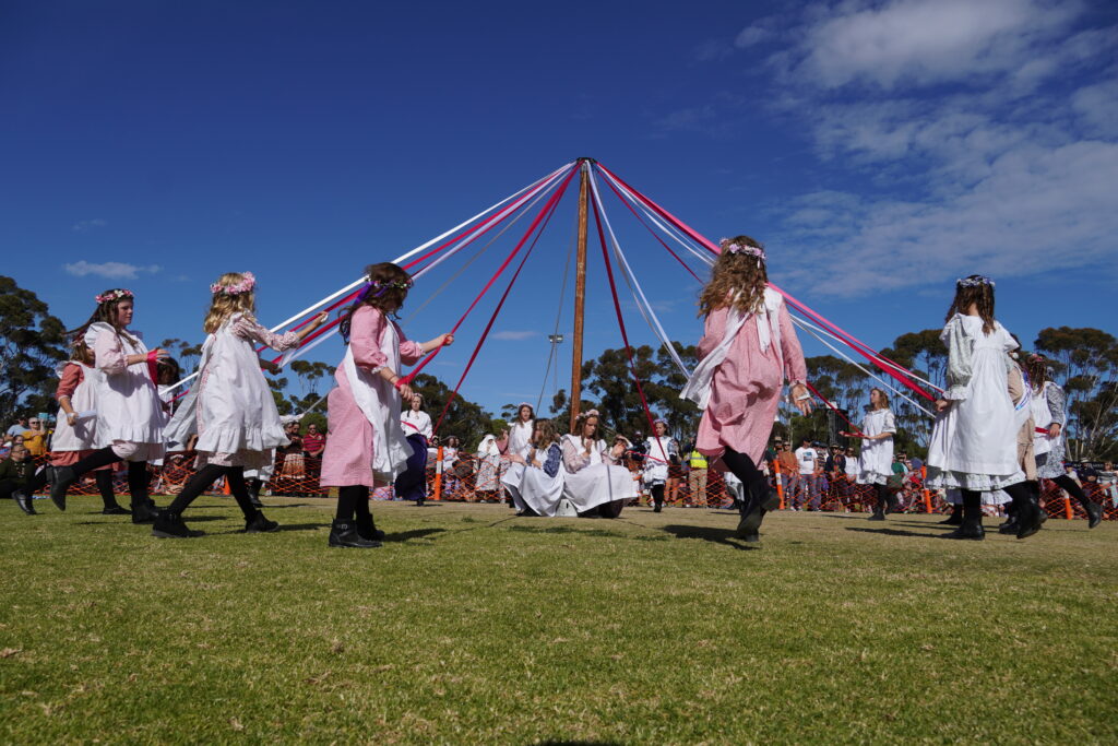 Girls dance around a maypole at the Kernewek Lowender 2023 festival parade on ther Copper Coast in Australia. The sky is blue and they are dressed in pink and white as they dance