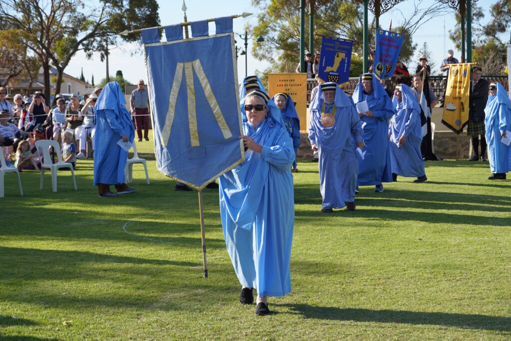A woman dressed in blue robes holds a blue and yellow flag at the Kernewek Lowender 2023 festival parade on the Copper Coast in Australia.  Other people follow, all dressed in traditional Cornish garb