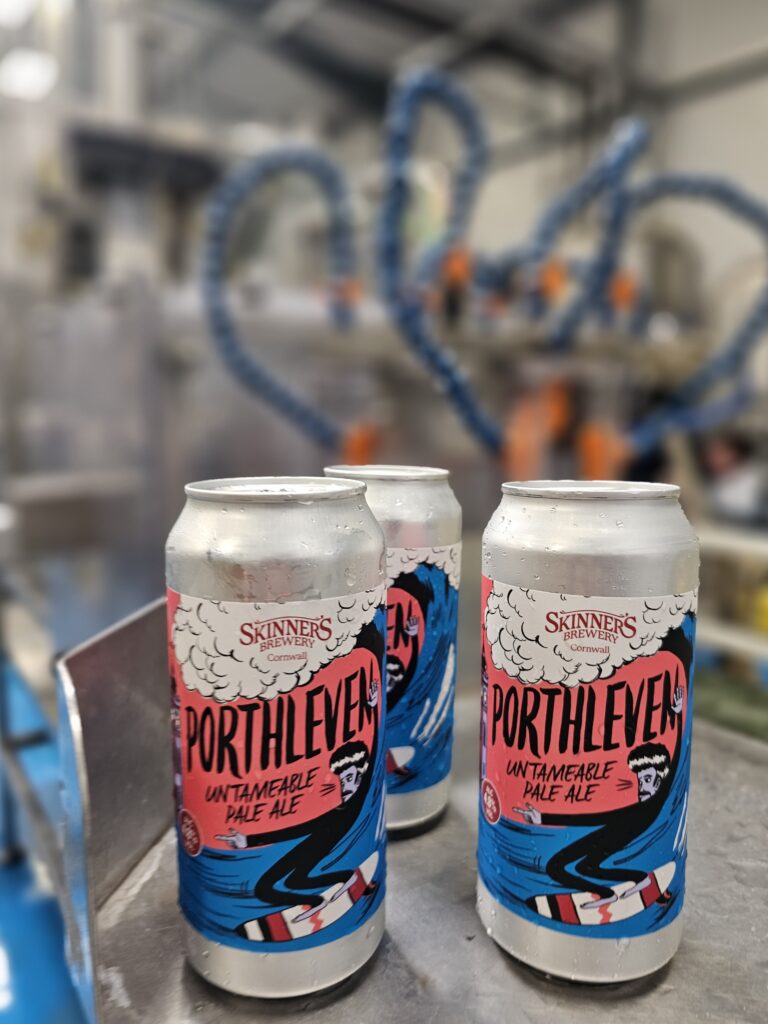 Cans of Porthleven beer by Skinner's Brewery sit on a table with the brewery machines in the background. This beer is being manufactured again by Goodh Brewery Company in partnership with Skinner's Brewery from June 2023
