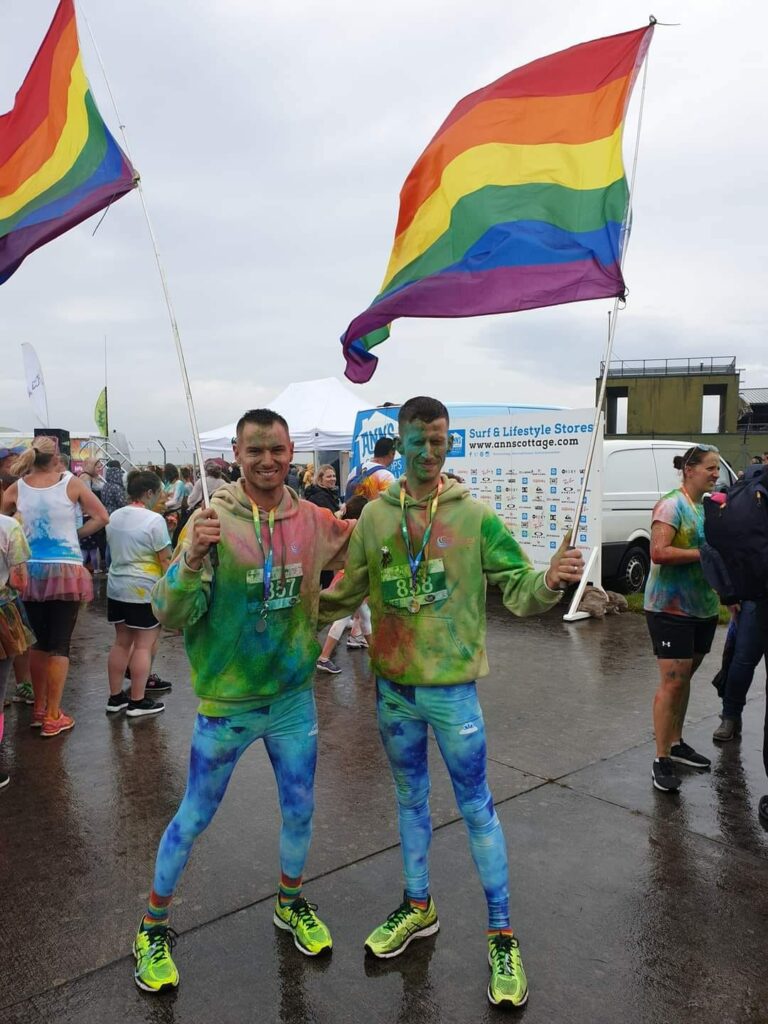 Two big rainbow flags are held aloft by Matthew Kenworthy Gomes, the chief executive of Cornwall Pride 2023, and his partner. They are both covered in green paint and stand next to the sea in Cornwall