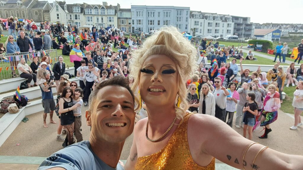 Matthew Kenworthy Gomes, Cornwall Pride chief executive, poses with an acclaimed drag artist in Cornwall during Cornwall Pride 2023. There are crowds of people standing and watching behind them