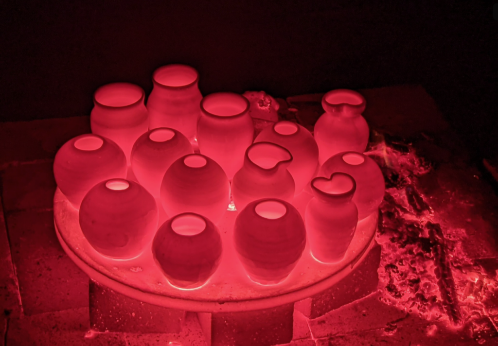 Red pottery looks almost translucent in the dark as it cools after being fired in the kiln at Swordfest 2023. There is an eerie glow to the scene