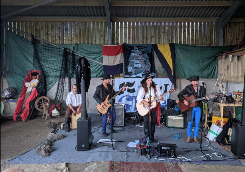 A band plays in a barn at Swordfest 2023 in Cornwall. There are banners and traditional items dotted all around