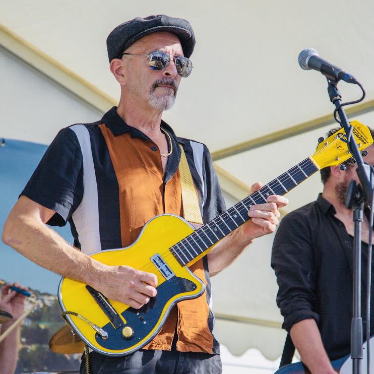 Plymouth blues guitarist Vince Lee plays a yellow guitar on stage ahead of the Looe Weekender 2023 festival in Cornwall. He has cool shades on and a flat cap and just looks like the personification of cool