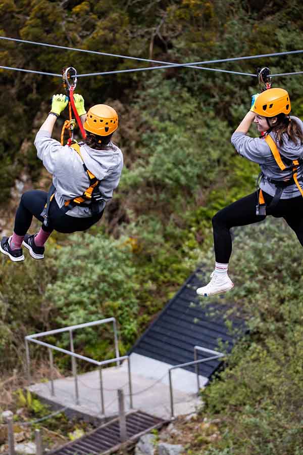 Two ladies in safety harnesses and hard hats are far above the quarry floor during the Zip Wire Safari at Via Ferrata Cornwall in Penryn. There is greenery, a fence and a path on the quarry floor. This is part of both the Zip Line Safari and Proper Cornwall's Summer Competition 2023