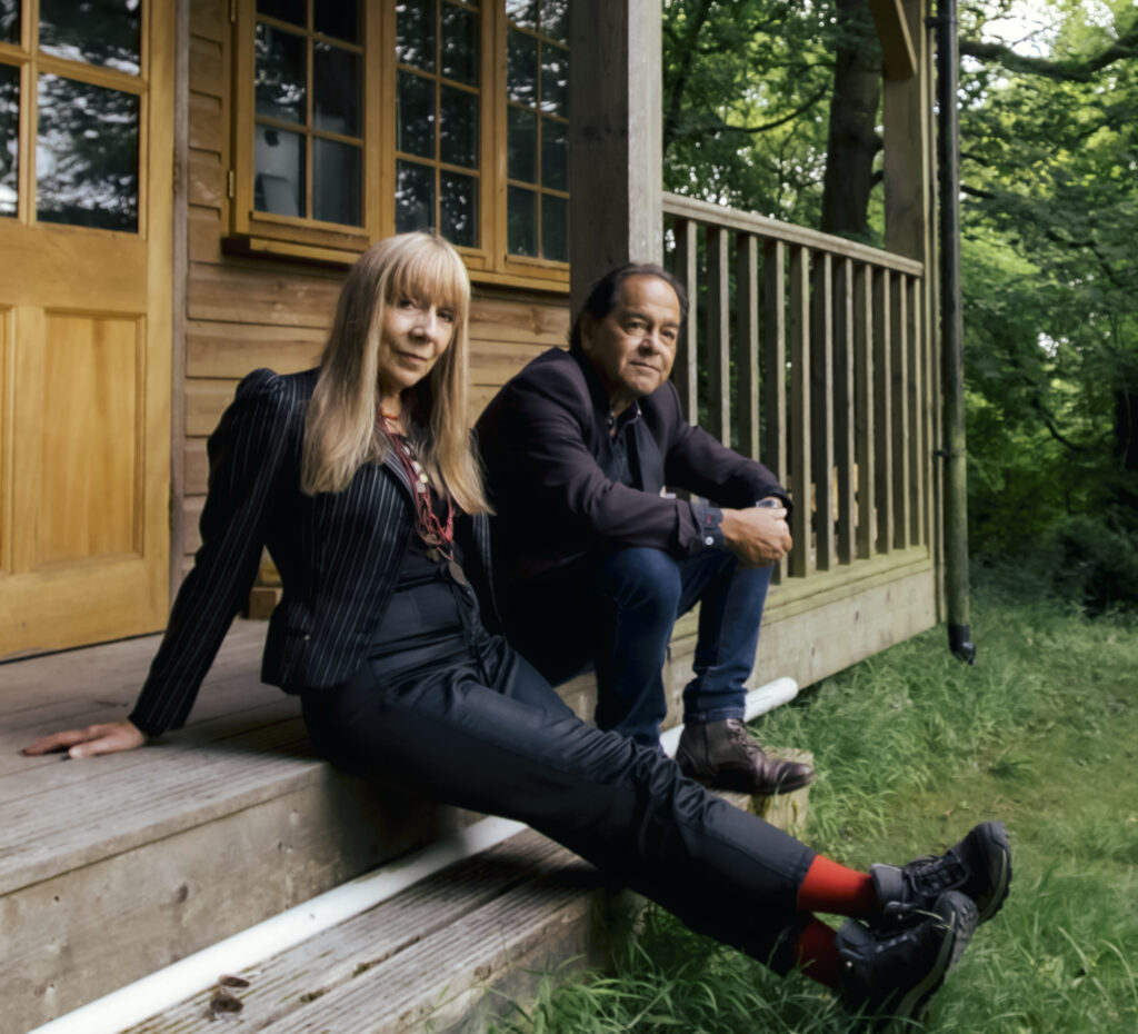Singer Charlie Dore and musician Julian Littman relax on the steps of a wooden lodge next to lush greenery. She has long blond hair and looks at us and he has a suit jacket on and looks into the distance. The duo perform at the North Cornwall Book Festival 2023 in St Endellion on Saturday 23 September 2023
