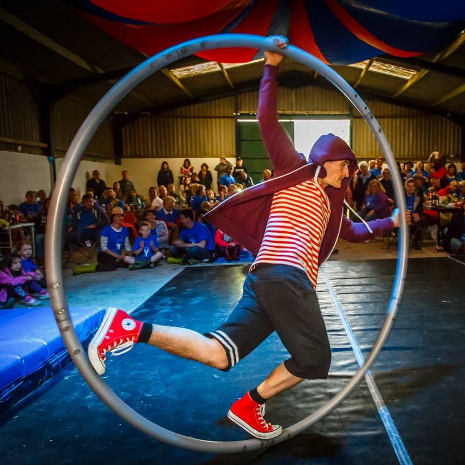 A member of Swamp Circus in Cornwall in a safety hat demonstrates a Cyr wheel on a mat in front of a crowd indoors. This is ahead of the Cornwall Circus Camp 2023 in August 2023