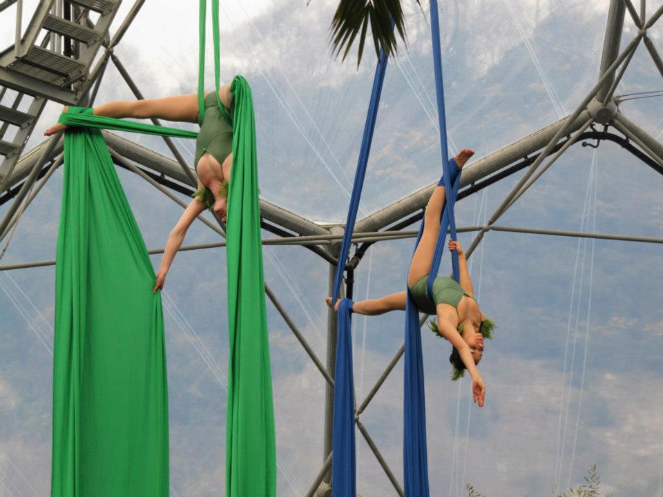 Two women in green and blue sashes dangle from the roof of the Eden Project in Cornwall as they show off their Swamp Circus skills. This is ahead of the Cornwall Circus Camp 2023 near Falmouth in August 2023