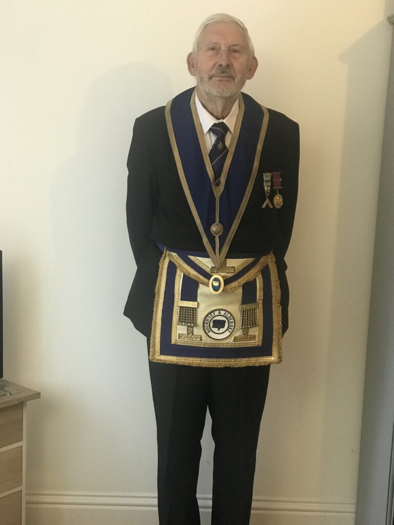 Mike Aireton, St Euny Lodge Master at Redruth Masonic Hall, stands in his apron and ceremonial suit against a blank wall. He looks at us and has medals on his lapel above his golden apron. This is ahead of his interview about the Cornwall Freemasons and masonic secrets with Proper Cornwall