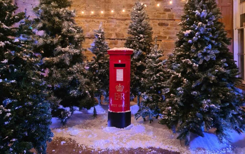 A red letterbox is surrounded by fir trees and snow inside Enys House near Penryn in Cornwall for the Enys Santa's Grotto 2023 experience. A stone wall from the manor house is behind and there are fairy lights above