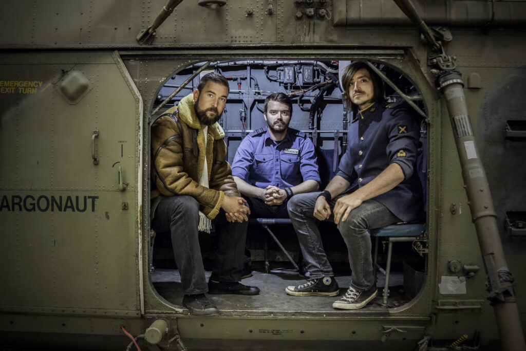 Heavy rock act Skam sit side-by-side in a military helicopter looking out at us. The trio play Cornwall Rocks 2024 in Tencreek Holiday Park near Looe. The lads look cool in military gear