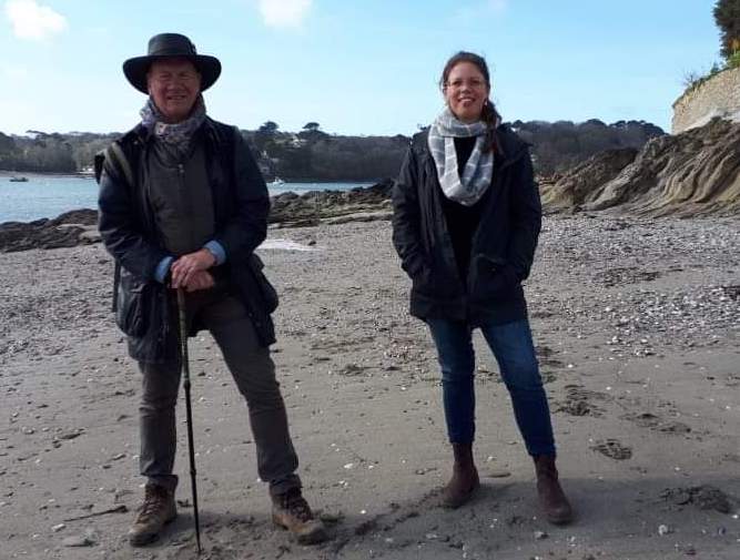 British politician and TV personality Michael Portillo stands next to The Cornish Bird, Elizabeth Dale, on a beach in Cornwall, with the sea in the background. Portillo wears a hat and holds a walking stick and Dale wears a scarf. Dale is quoted in Lawrence McNeela's story on the renaissance of Cornish folklore in modern times, published by Proper Cornwall in February 2024
