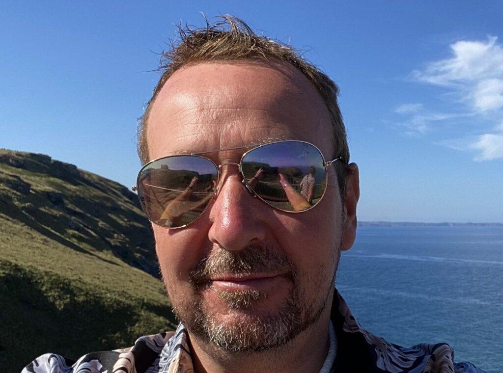 Cornwall author Lawrence McNeela wears sunglasses and stands in front of a Cornish cliff and the sea. He guest authors articles for Proper Cornwall. The sky is blue and beautiful